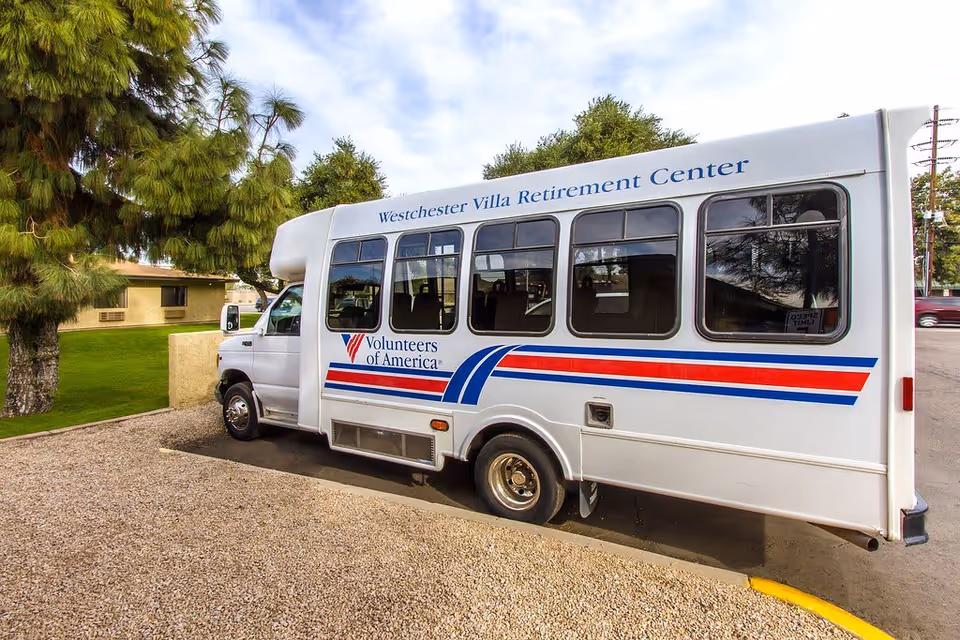 A white Volunteers of America shuttle bus labeled Westchester Villa Retirement Center parked outside near trees and a low building.