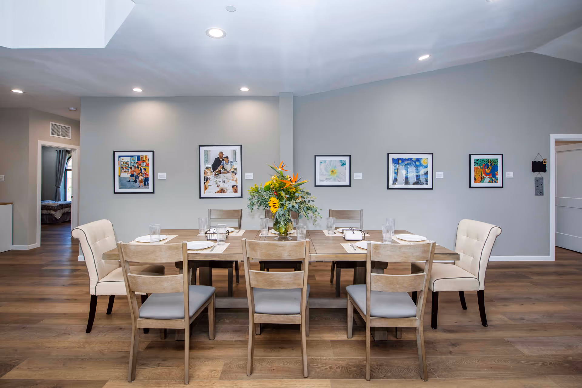 A dining room with a wooden table set for six people, including plates, glasses, and napkins. The table has a centerpiece with a vase of colorful flowers. The room has light gray walls adorned with five framed artworks. The floor is wooden, and there are two upholstered chairs at the ends of the table and four wooden chairs along the sides. Two doorways are visible, one leading to a bedroom and the other to another room.