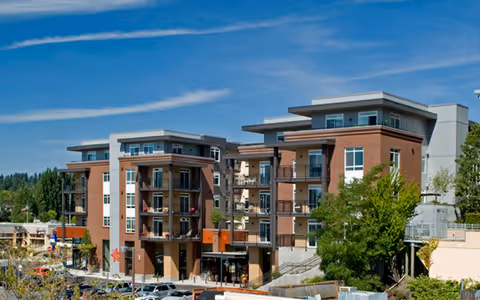 Exterior view of a modern multi-story residential building with balconies, surrounded by trees and parked cars under a blue sky with wispy clouds.