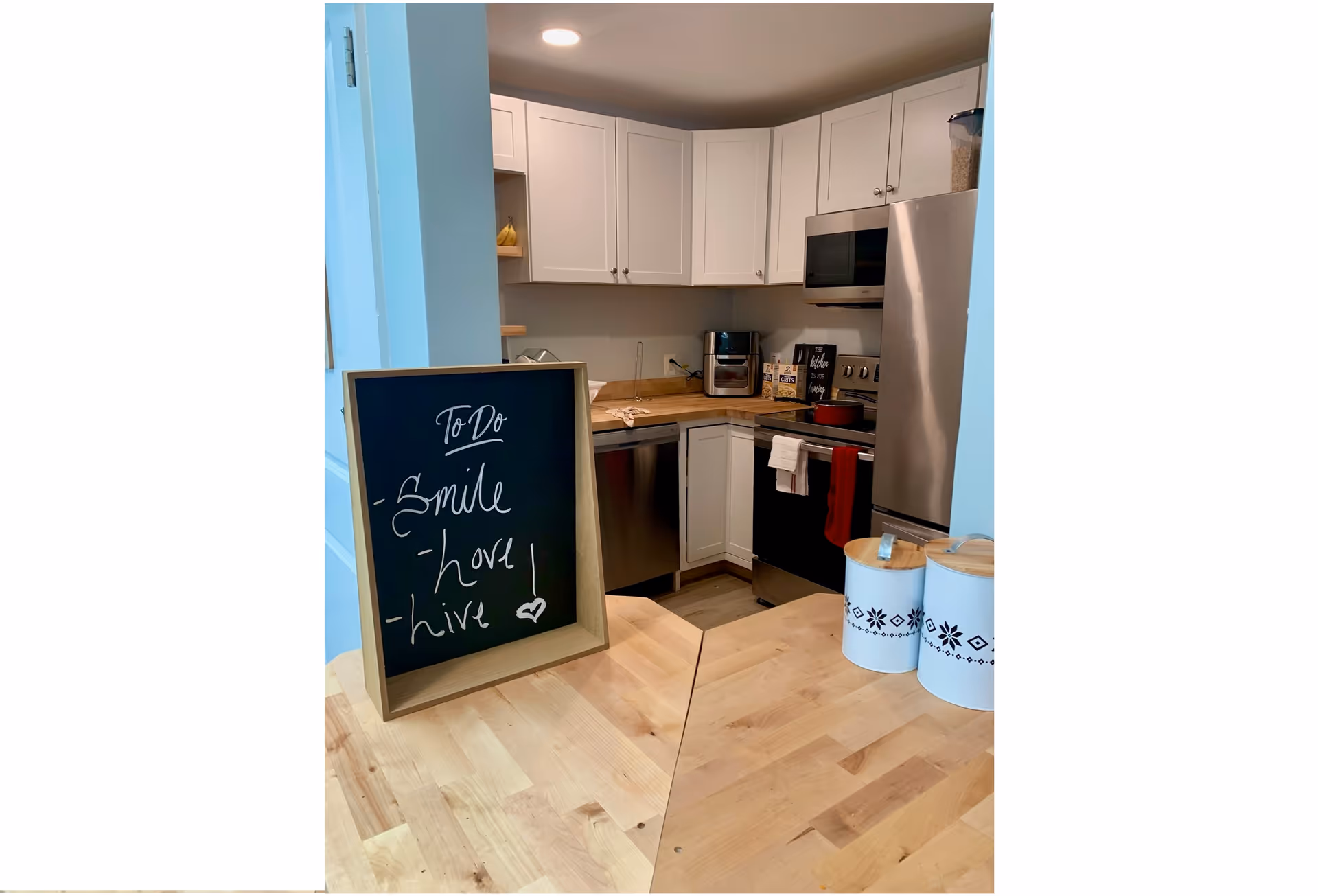 Compact kitchen with white cabinets, stainless-steel appliances, a wooden countertop island, and a framed chalkboard with a handwritten to-do list.