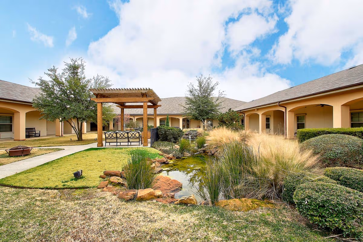 Outdoor courtyard area at The Oaks At Flower Mound featuring a small pond with rocks and tall grasses, a wooden pergola with seating underneath, surrounded by a single-story building with arched walkways and neatly trimmed bushes and trees under a partly cloudy sky.