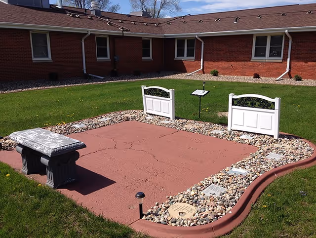 A serene garden area featuring a stone bench, decorative stones, and white gates, surrounded by green grass and a brick building in the background.