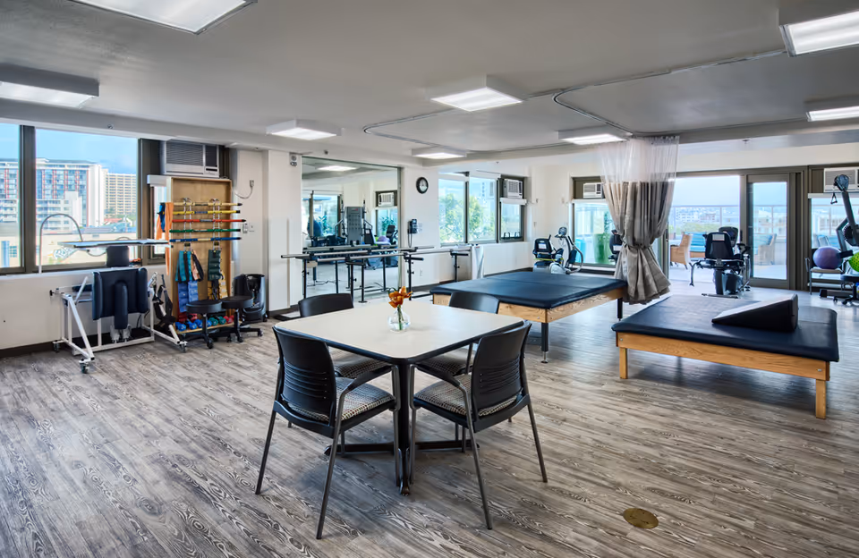 Spacious interior therapy and fitness room with treatment tables, exercise equipment, and a small seating table by large windows.