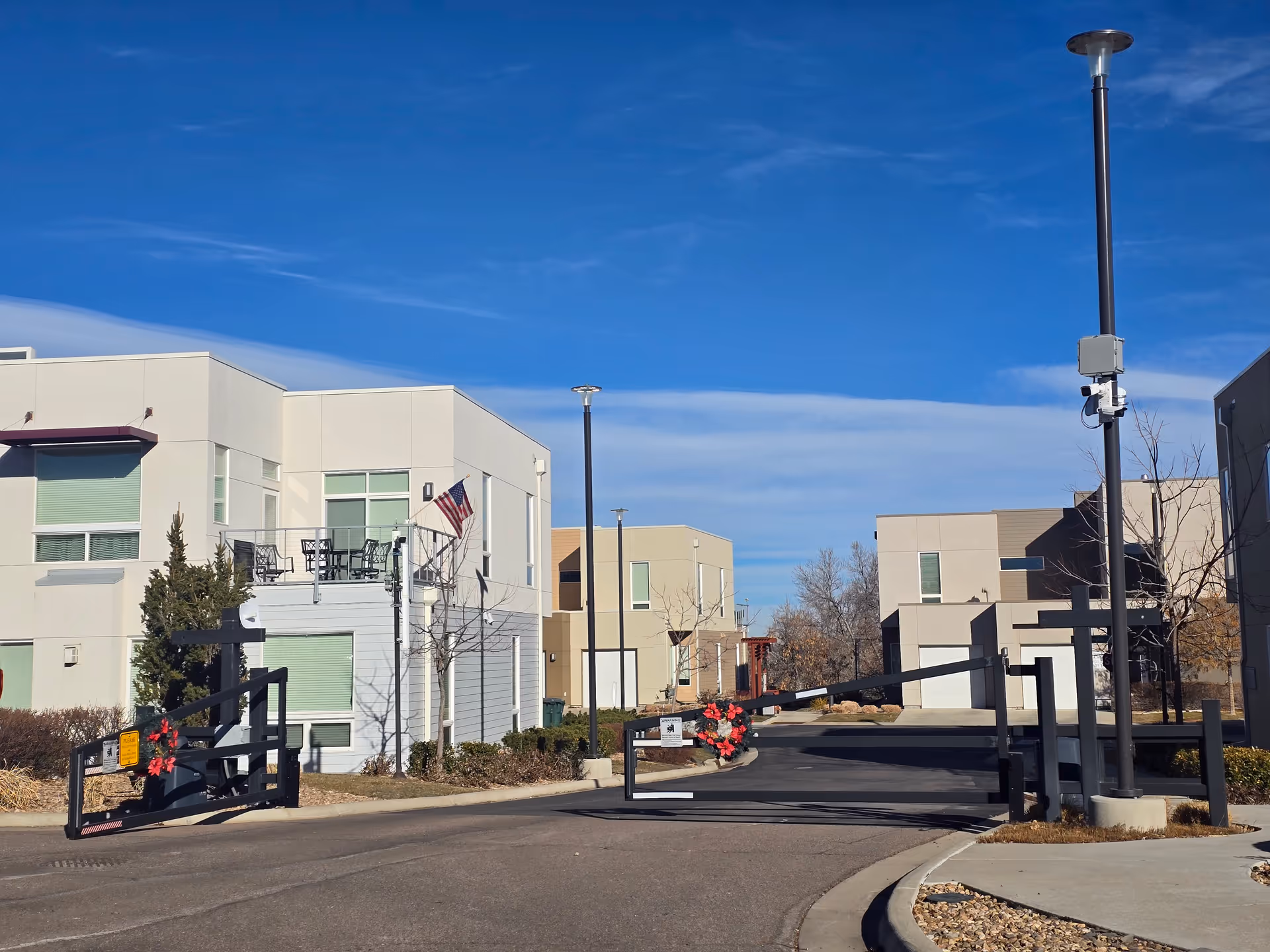 Gated entrance to a modern low-rise residential complex with beige and white buildings, an American flag, and a clear blue sky.