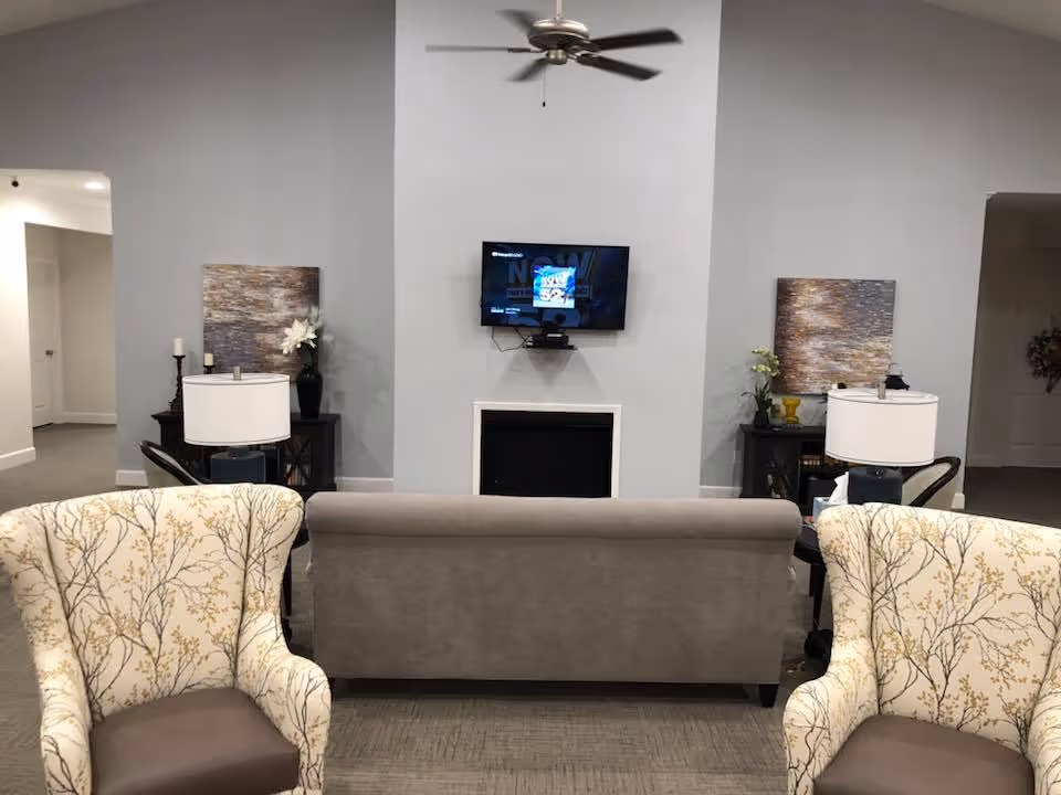 A living room area with a gray sofa facing a wall-mounted TV above a fireplace. Two patterned armchairs with floral designs are positioned in the foreground. On either side of the fireplace are small black tables with lamps, decorative items, and artwork hanging on the wall. The walls are painted light gray and the floor is carpeted.