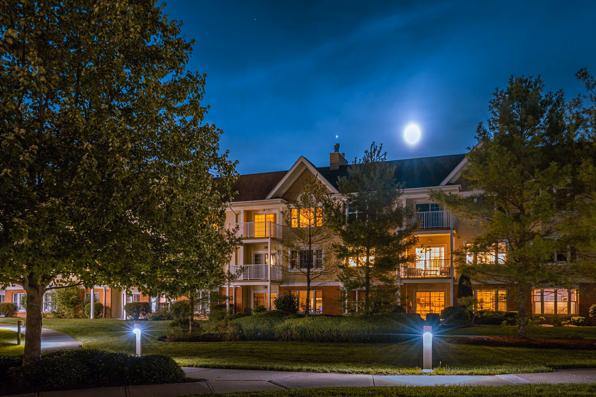 Nighttime exterior view of a multi-story residential building with balconies and lit windows, surrounded by trees and well-maintained landscaping under a clear sky with a bright moon.