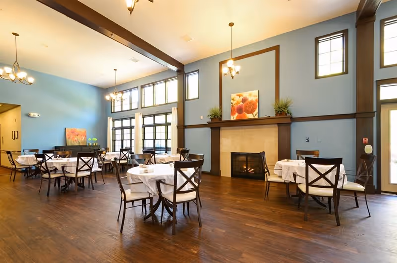 A spacious dining room with several round tables covered with white tablecloths and surrounded by wooden chairs with cushioned seats. The room features large windows allowing natural light, blue walls with dark wood trim, a fireplace with a floral painting above it, and hanging light fixtures.