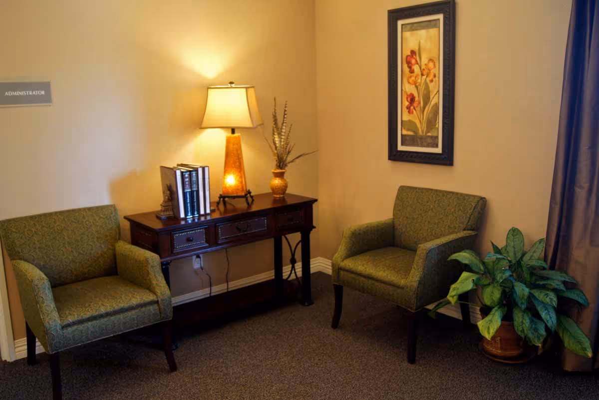 A cozy office waiting area with two green upholstered armchairs, a wooden console table with a lit table lamp, books, and a decorative vase. A framed floral artwork hangs on the wall above one chair, and a potted plant is placed near a window with curtains.