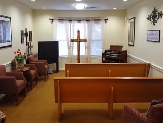 A small chapel room with wooden pews facing a wooden cross in front of a window with sheer white curtains. To the left are several upholstered chairs and a TV, and to the right is an organ. The walls are decorated with framed pictures and a decorative metal wall hanging.