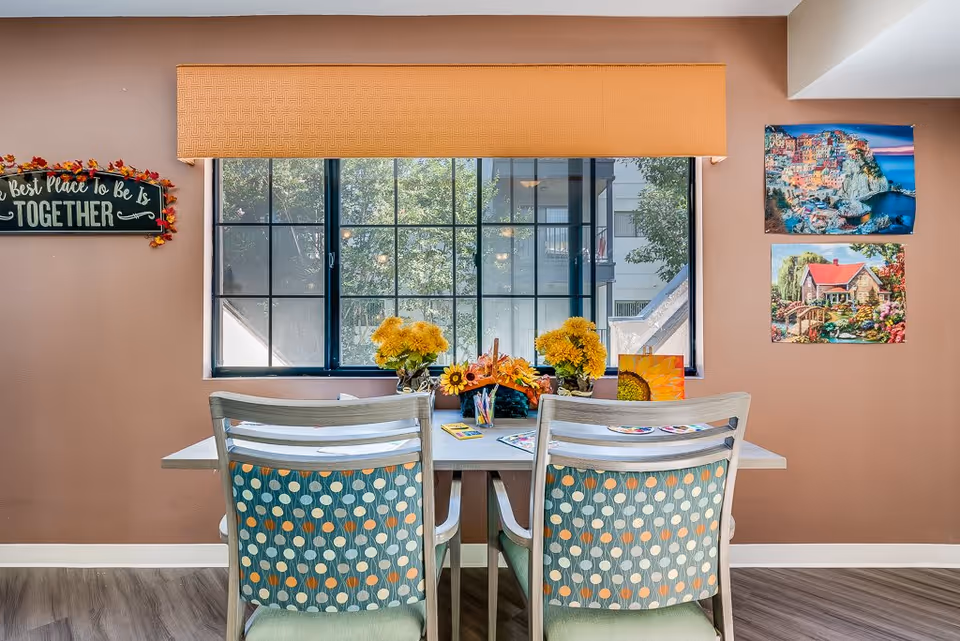 A cozy interior room with a table and two chairs facing a large window. The table is decorated with yellow flowers, art supplies, and colorful paintings. The walls are painted light brown with a sign that reads 'The Best Place To Be Is TOGETHER' on the left and two landscape paintings on the right.
