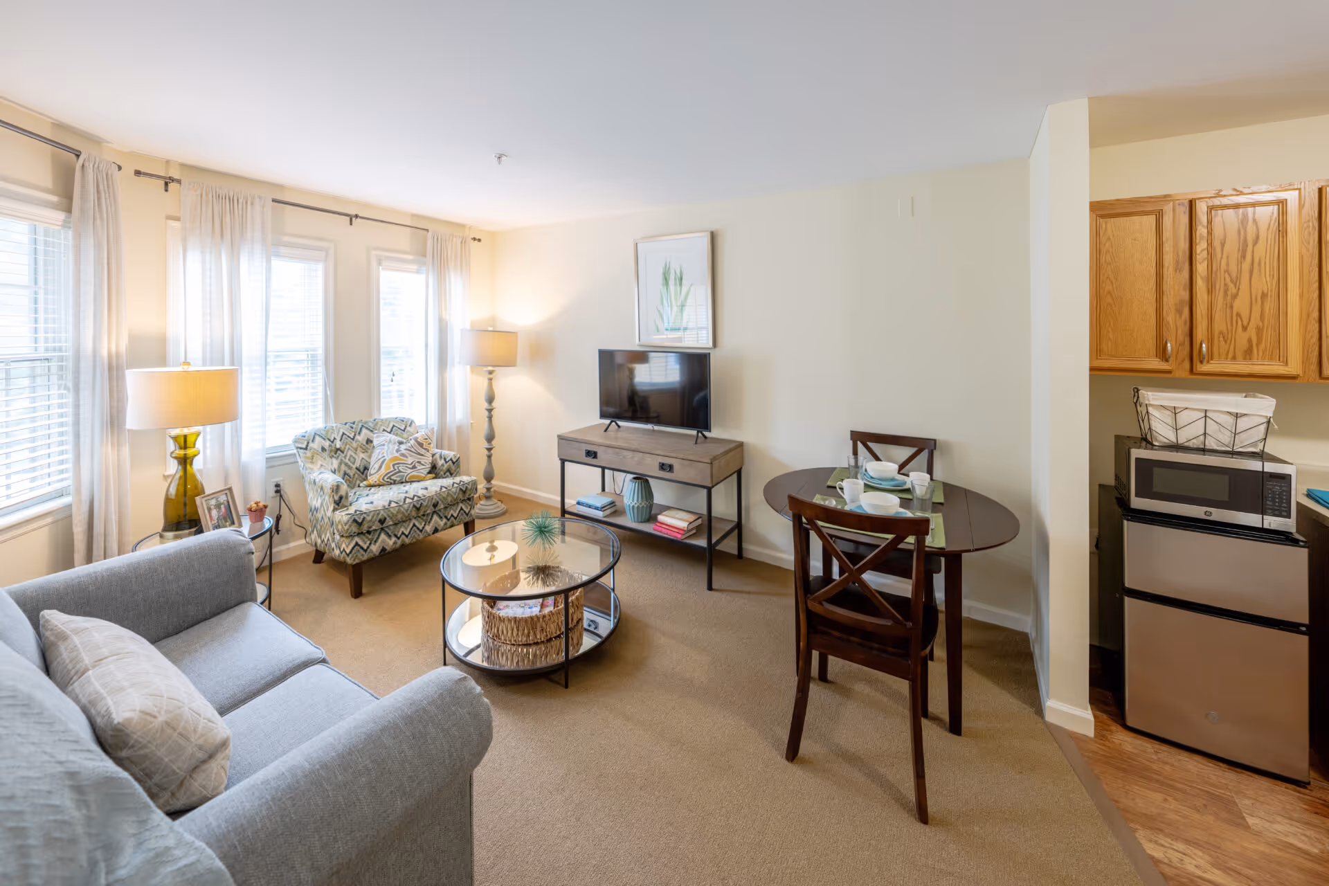 Bright furnished living room with a sofa, patterned armchair, round coffee table, TV on a console, small dining table and a kitchenette area.