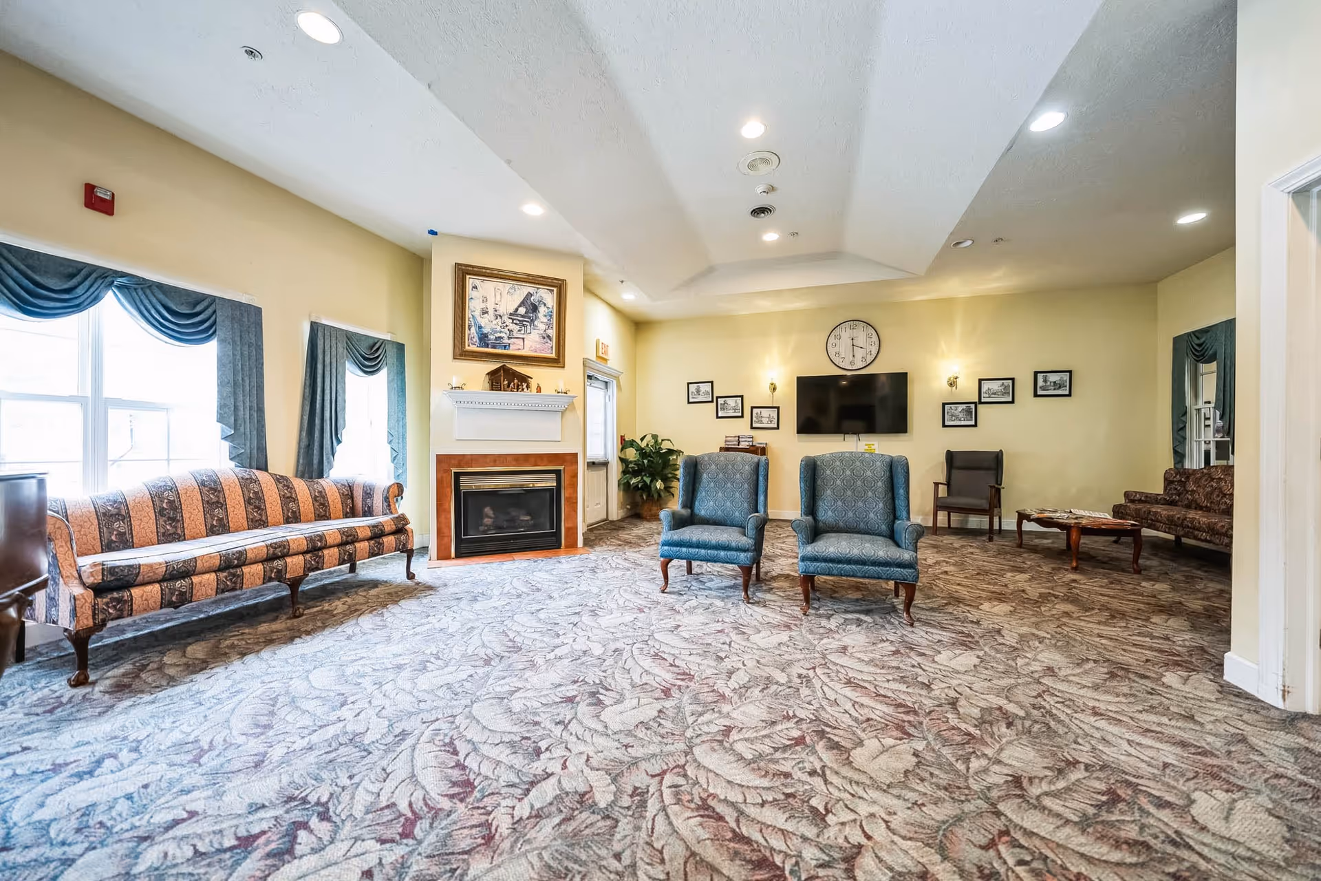 A spacious senior living facility common area with patterned carpet, two blue upholstered armchairs, a patterned sofa near windows with blue curtains, a fireplace with a framed picture above it, a wall-mounted TV, a clock, and several framed pictures on the wall.