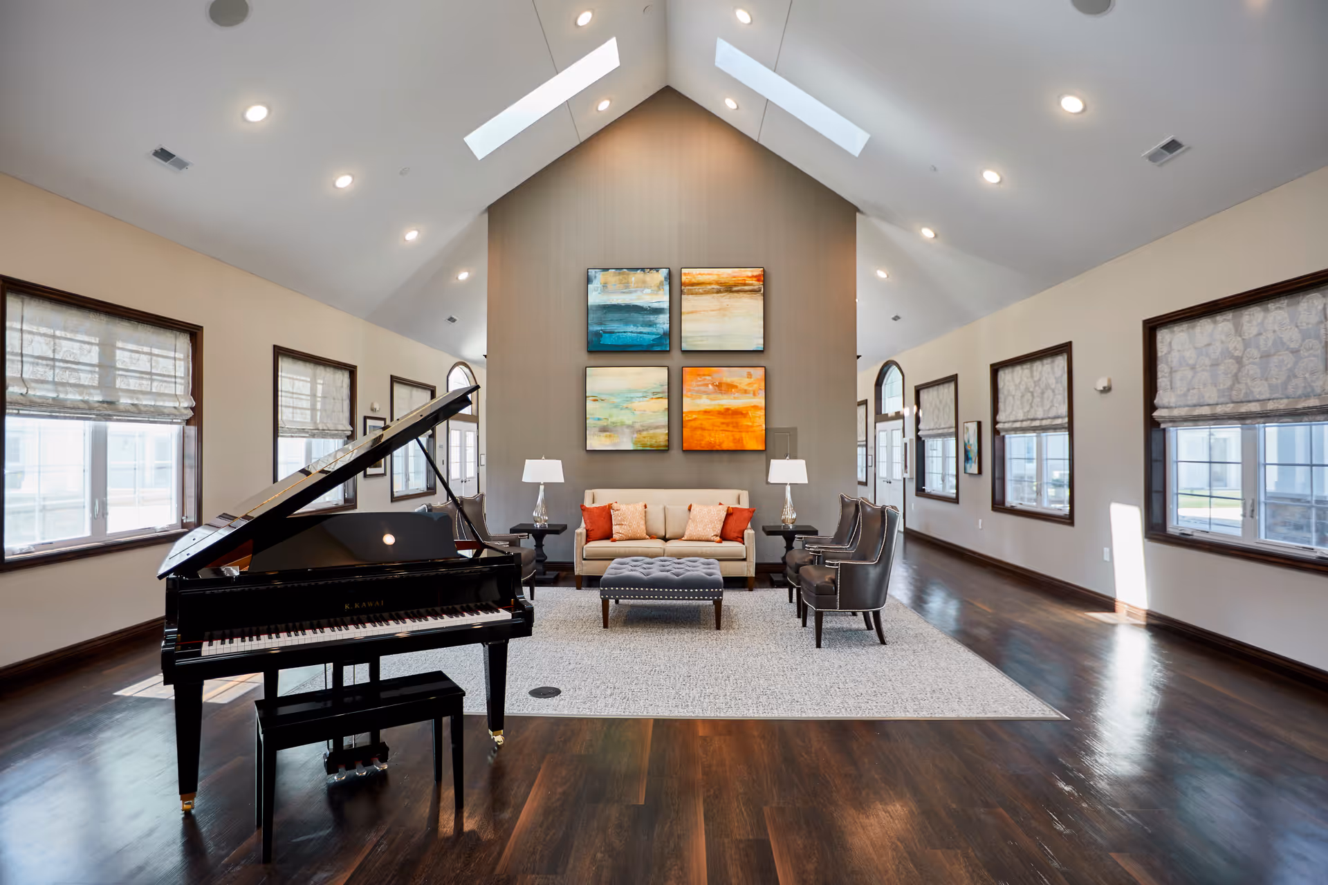 A spacious, well-lit living room with a high vaulted ceiling featuring skylights. The room has dark hardwood floors and multiple windows with patterned Roman shades. A black grand piano with a matching bench is positioned on the left side. In the center, there is a beige sofa with orange and beige cushions, flanked by two side tables with lamps. Two dark leather armchairs and a tufted ottoman are arranged on a large area rug. Four colorful abstract paintings hang on the wall behind the sofa.