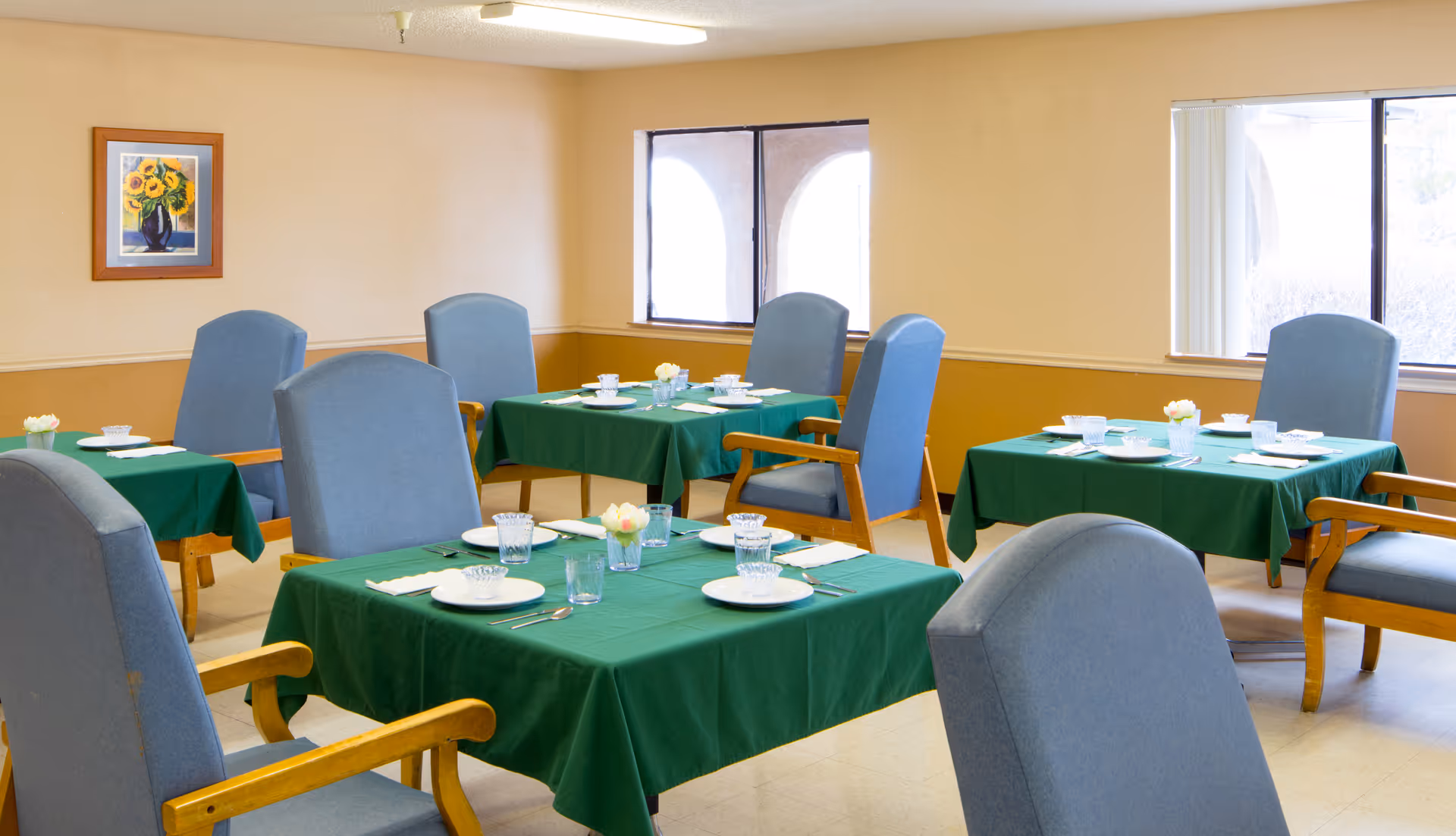 A dining room with several square tables covered with green tablecloths, each set with white plates, glasses, utensils, and small flower arrangements. The room has light yellow walls, large windows letting in natural light, and blue cushioned chairs with wooden armrests around the tables.
