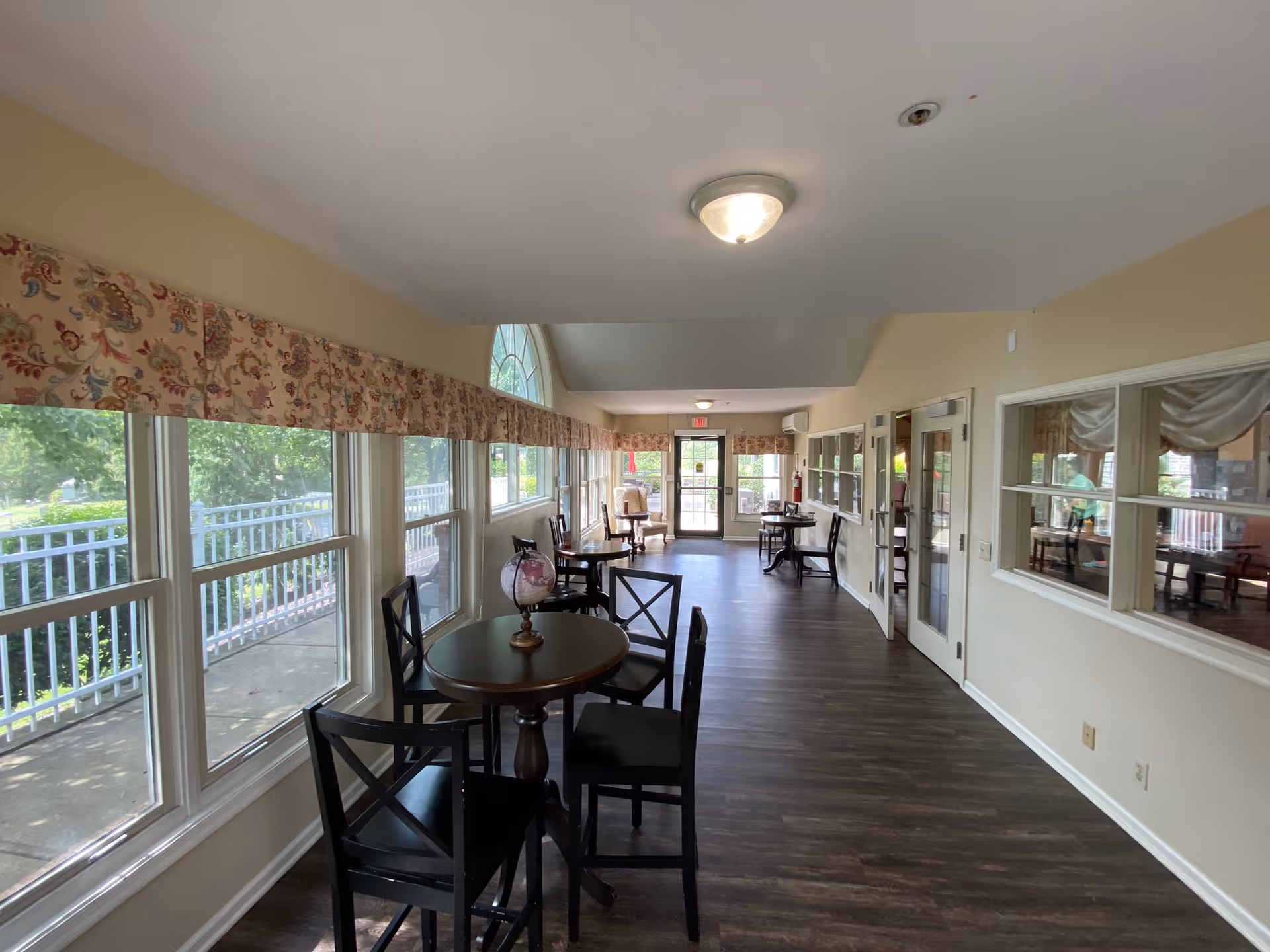 A bright interior hallway with large windows on the left side covered by floral valances, letting in natural light. There are several small round tables with black chairs arranged along the hallway. The floor is dark wood, and the walls are painted light beige. At the far end, there is a glass door leading outside, and on the right side, there are windows looking into another room.