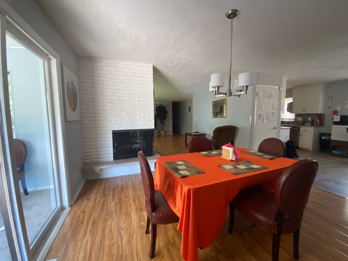 Dining room with red tablecloth, chairs, a brick fireplace, and visible kitchen.