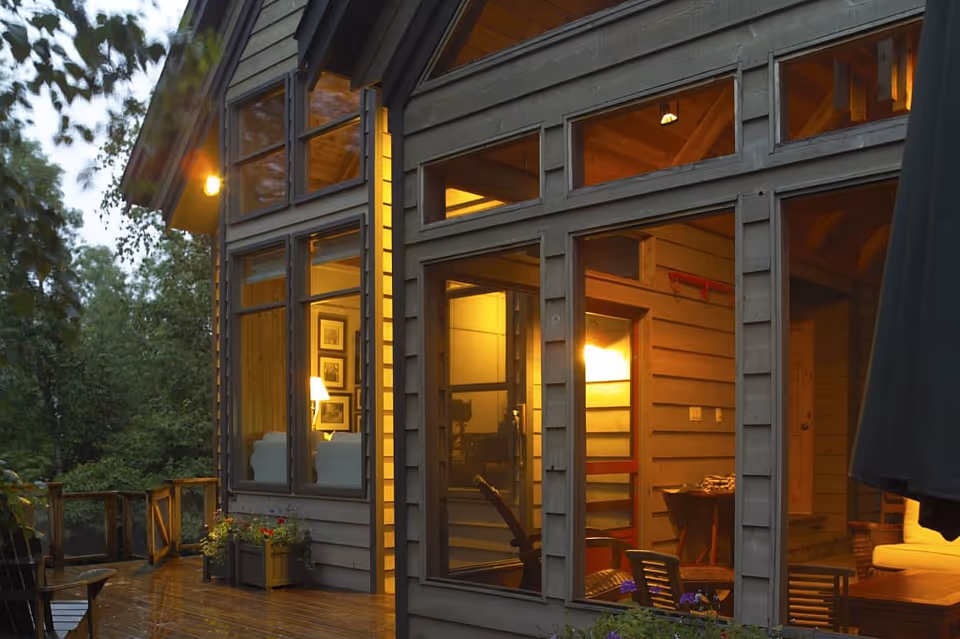 Exterior view of a wooden house at dusk with large windows showing a warmly lit interior. The house has a wooden deck with some outdoor furniture and plants, surrounded by trees.