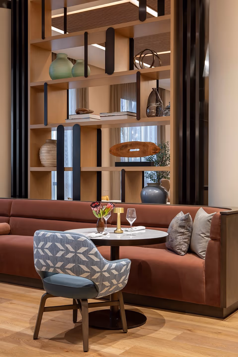 A cozy seating area in a senior living facility featuring a round table set with a small vase of flowers, a candle holder, and two wine glasses. The table is surrounded by a patterned blue armchair and a built-in rust-colored cushioned bench with decorative pillows. Behind the seating area is a wooden shelving unit with various decorative items including vases and books, and light wood flooring.