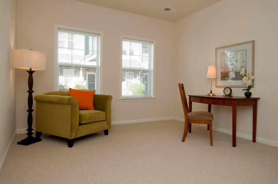 A simple, well-lit room with beige walls and carpet. There is a green armchair with an orange pillow on the left side next to a tall floor lamp. On the right side, there is a wooden desk with a chair, a small table lamp, a clock, and a potted orchid. Two windows with white blinds allow natural light into the room.
