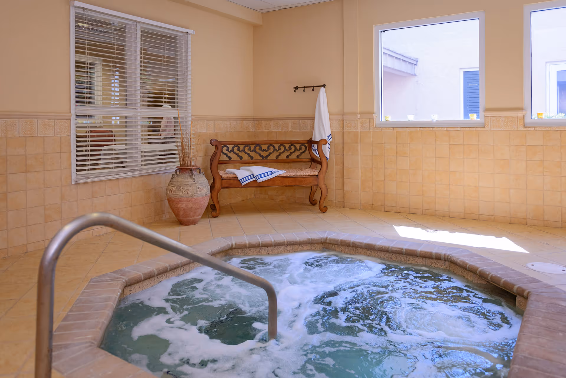 A tiled indoor whirlpool/jacuzzi with bubbling water, a wooden bench with towels, and windows letting in light.