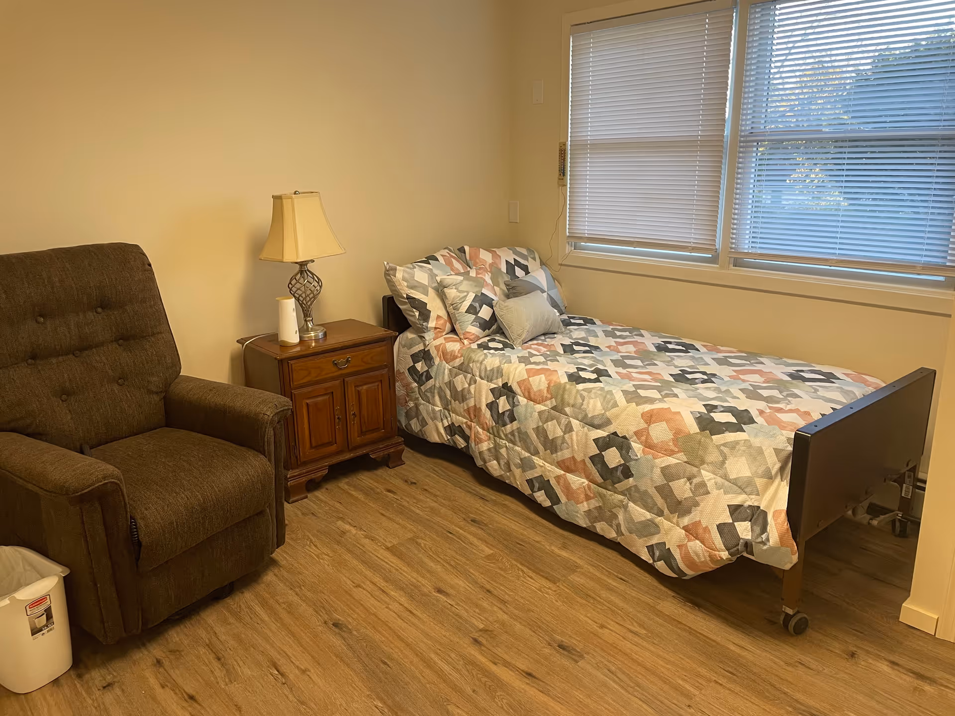 A small bedroom with a single bed covered in a patterned quilt, a wooden nightstand with a lamp and a white object on it, a brown upholstered recliner chair, and a window with closed blinds letting in some natural light.
