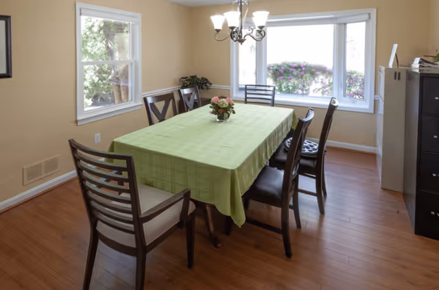 A dining room with a rectangular table covered by a green tablecloth, surrounded by six wooden chairs with cushions. A small flower arrangement is placed in the center of the table. The room has wooden flooring, beige walls, two windows with views of greenery outside, and a chandelier hanging above the table. There is a black cabinet on the right side of the room.
