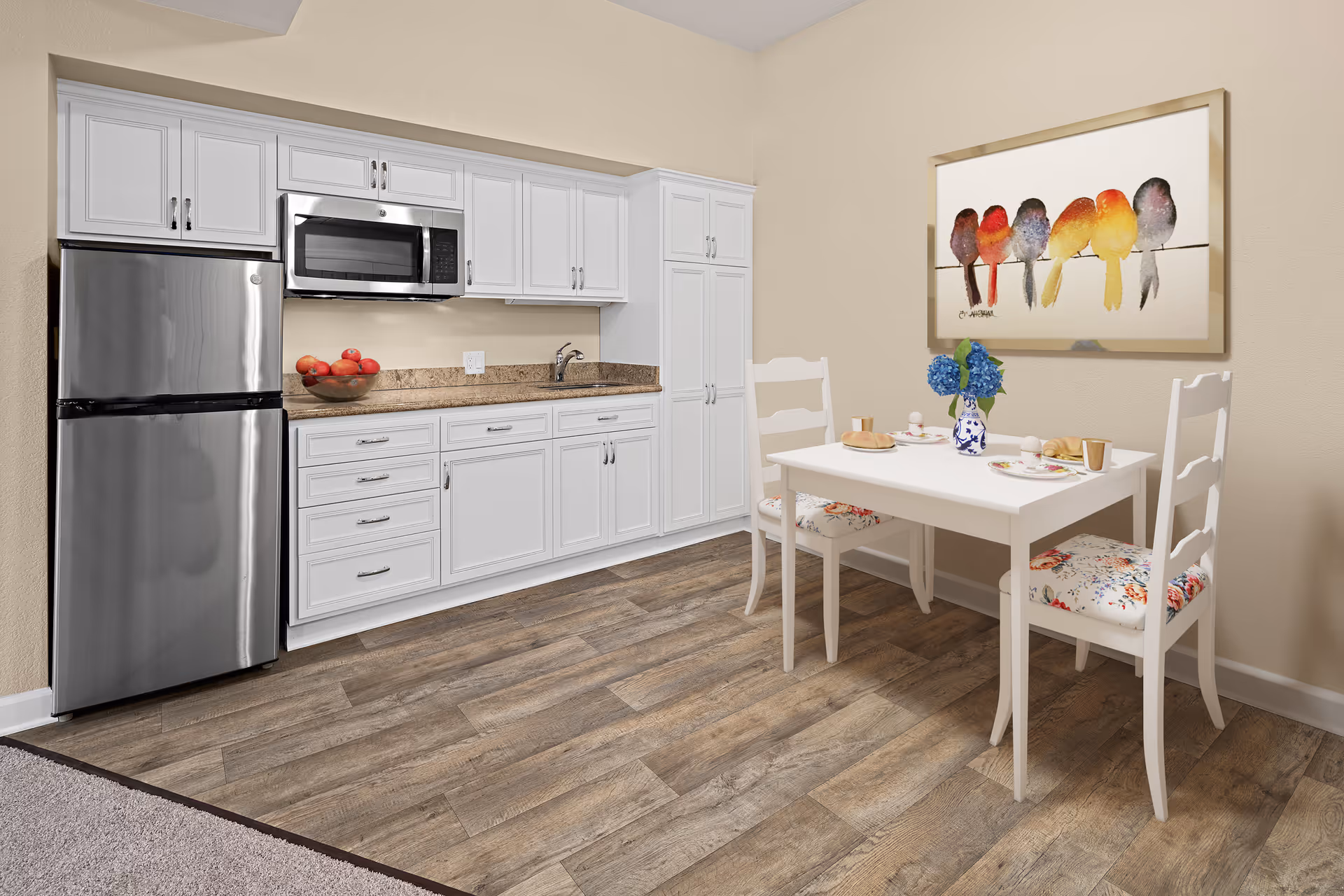 A small kitchen area with white cabinets, a stainless steel refrigerator, and a microwave above the countertop. Next to the kitchen is a white dining table set for two with floral cushioned chairs and a vase with blue flowers. A colorful painting of birds on a wire hangs on the wall above the table.