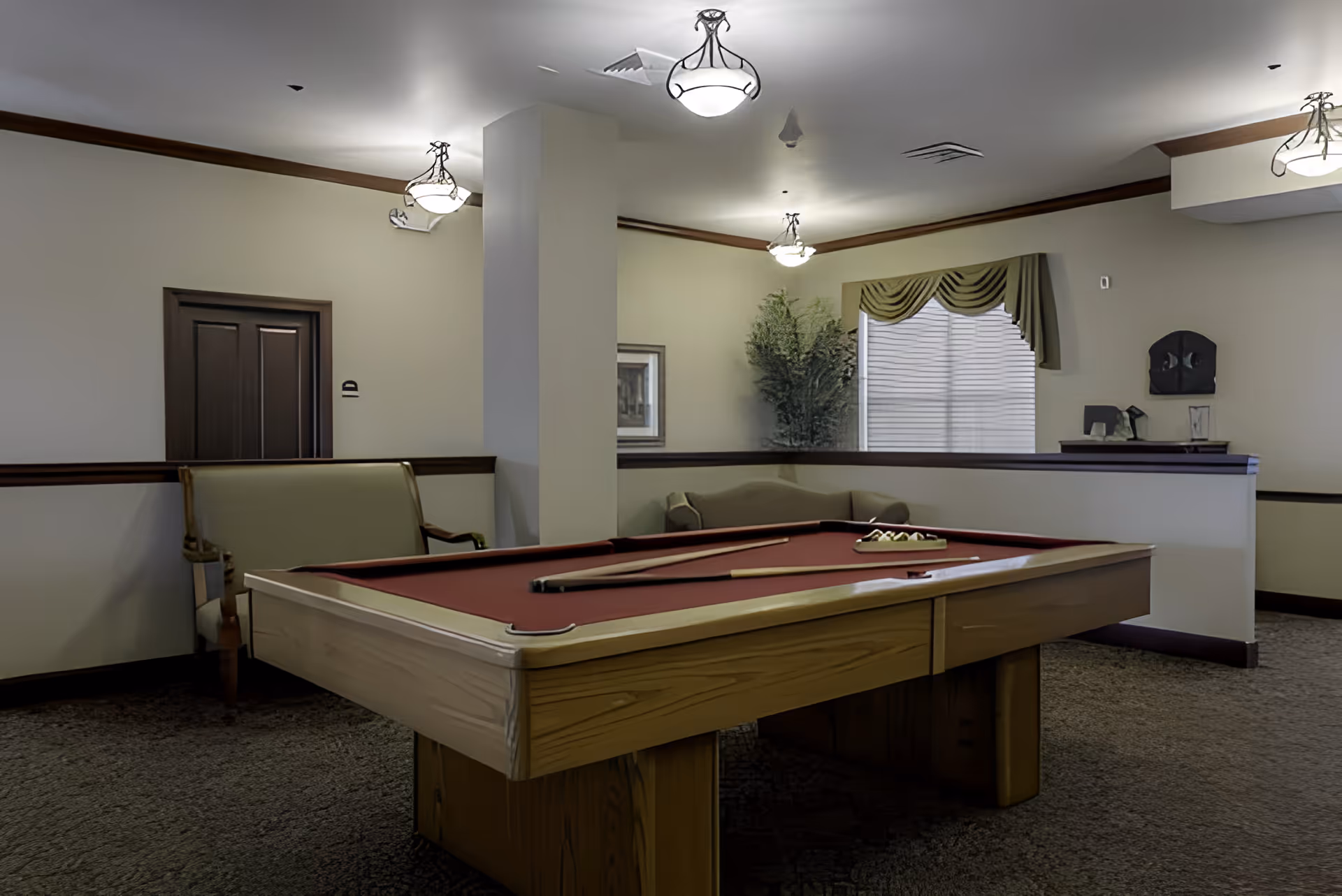 Interior room with a pool table featuring a red felt surface, two pool cues, and a rack of billiard balls. The room has beige walls with dark wood trim, carpeted floor, ceiling lights, a green upholstered bench, a green sofa, a window with green valance curtains, and a potted plant in the corner.