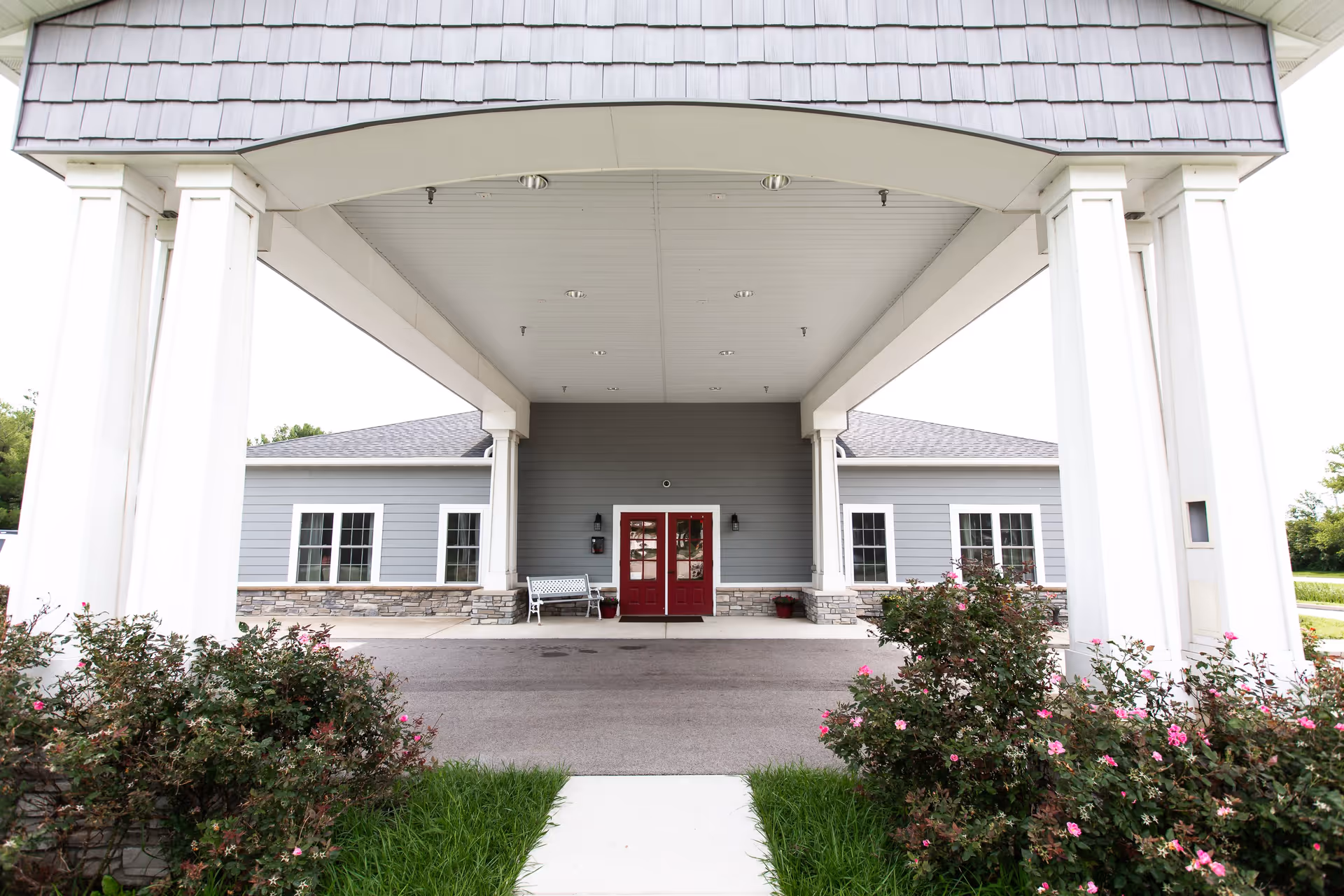 Entrance of a building with a covered driveway supported by white columns, gray siding, stone accents, red double doors, a white bench, and flowering bushes on either side of the walkway.