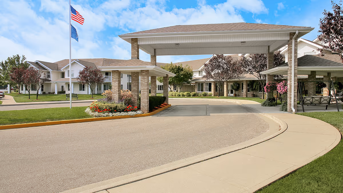 Exterior view of Independence Village of Columbus showing a covered entrance with brick pillars, landscaped flower beds, green lawns, trees, and two flagpoles with the American and state flags against a blue sky.