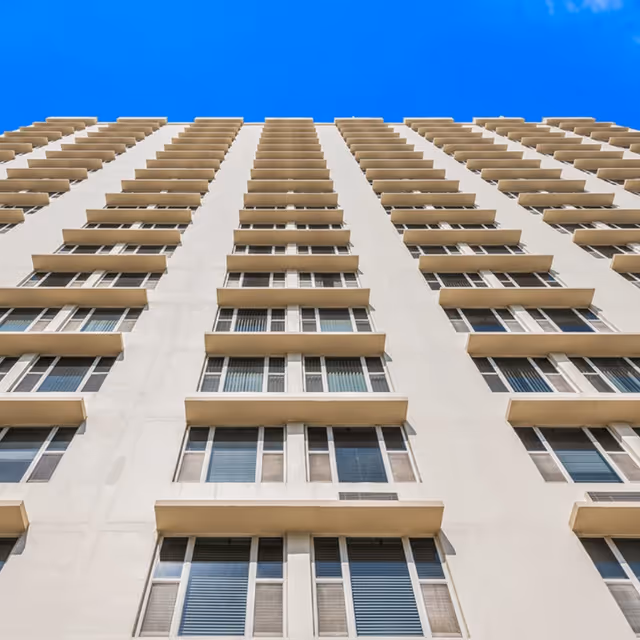Upward view of a tall residential building with multiple rows of windows and balconies under a clear blue sky.