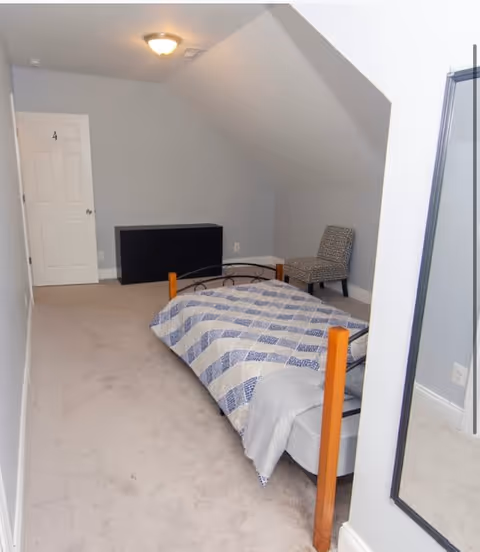 A small bedroom with a single bed covered in a blue and white checkered quilt. The room has light gray walls and beige carpet. There is a black chest of drawers against the far wall, a patterned chair in the corner, and a white door on the left side. A ceiling light fixture is visible, and a large mirror is mounted on the right wall.
