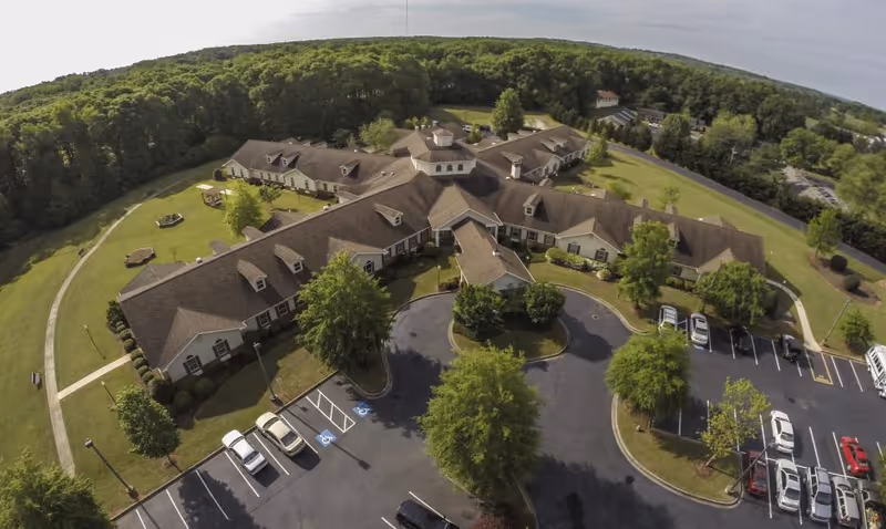 Aerial view of The Gables At Cobb Village senior living facility showing a large, single-story building with multiple wings surrounded by green lawns, trees, and a parking lot with several cars parked. The building is set against a backdrop of dense forest under a clear sky.