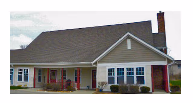 Exterior view of a single-story residential building with a gable roof, multiple windows, a covered porch with red pillars, and a chimney on the right side.