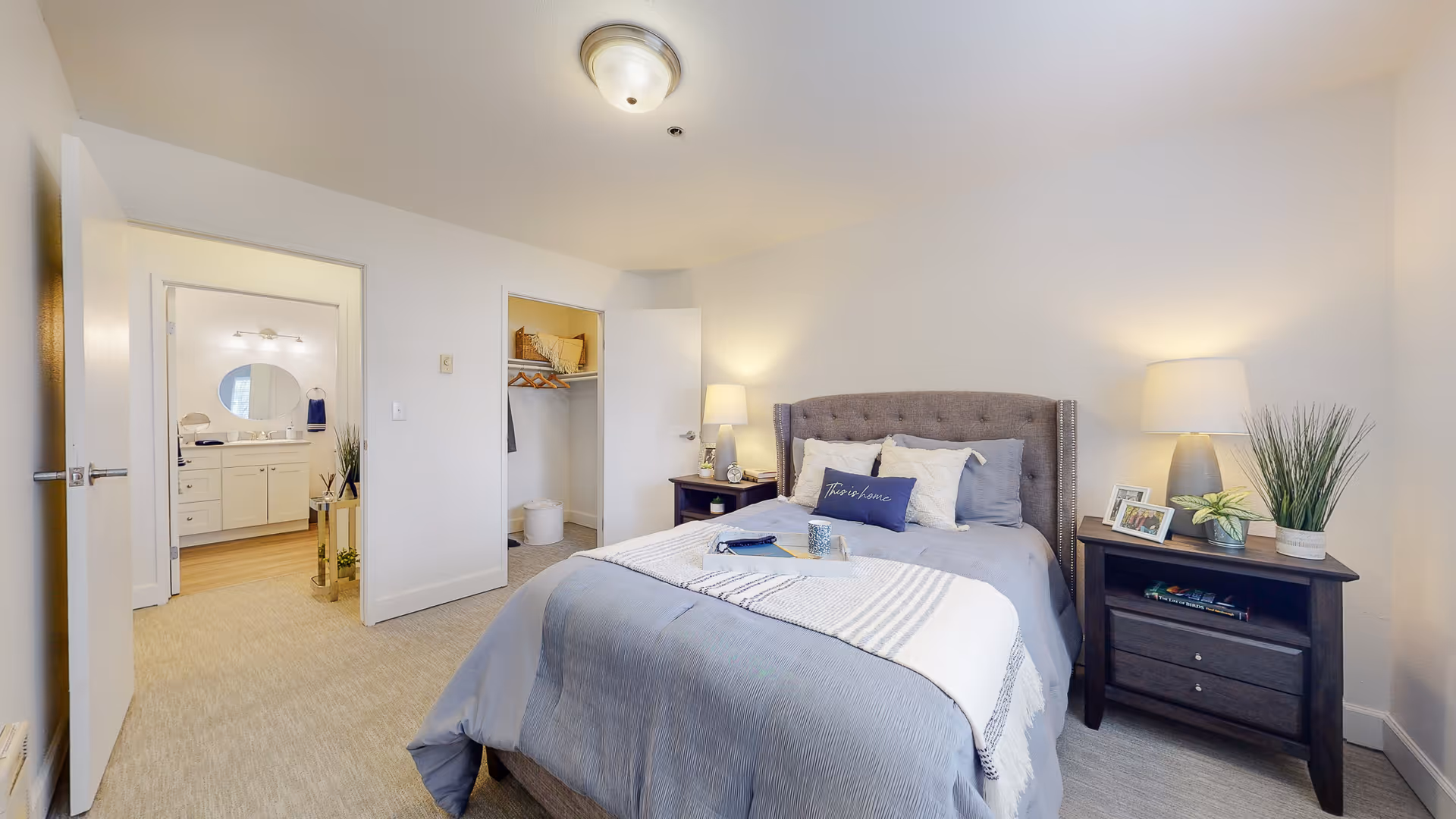 A neatly made bed with gray and white bedding in a bright bedroom. There are two dark wooden nightstands on either side of the bed, each with a lamp and decorative items including framed photos and plants. An open door reveals a walk-in closet with shelves and hanging clothes, and another door leads to a bathroom with a vanity and round mirror. The room has light-colored walls and carpeted floor.