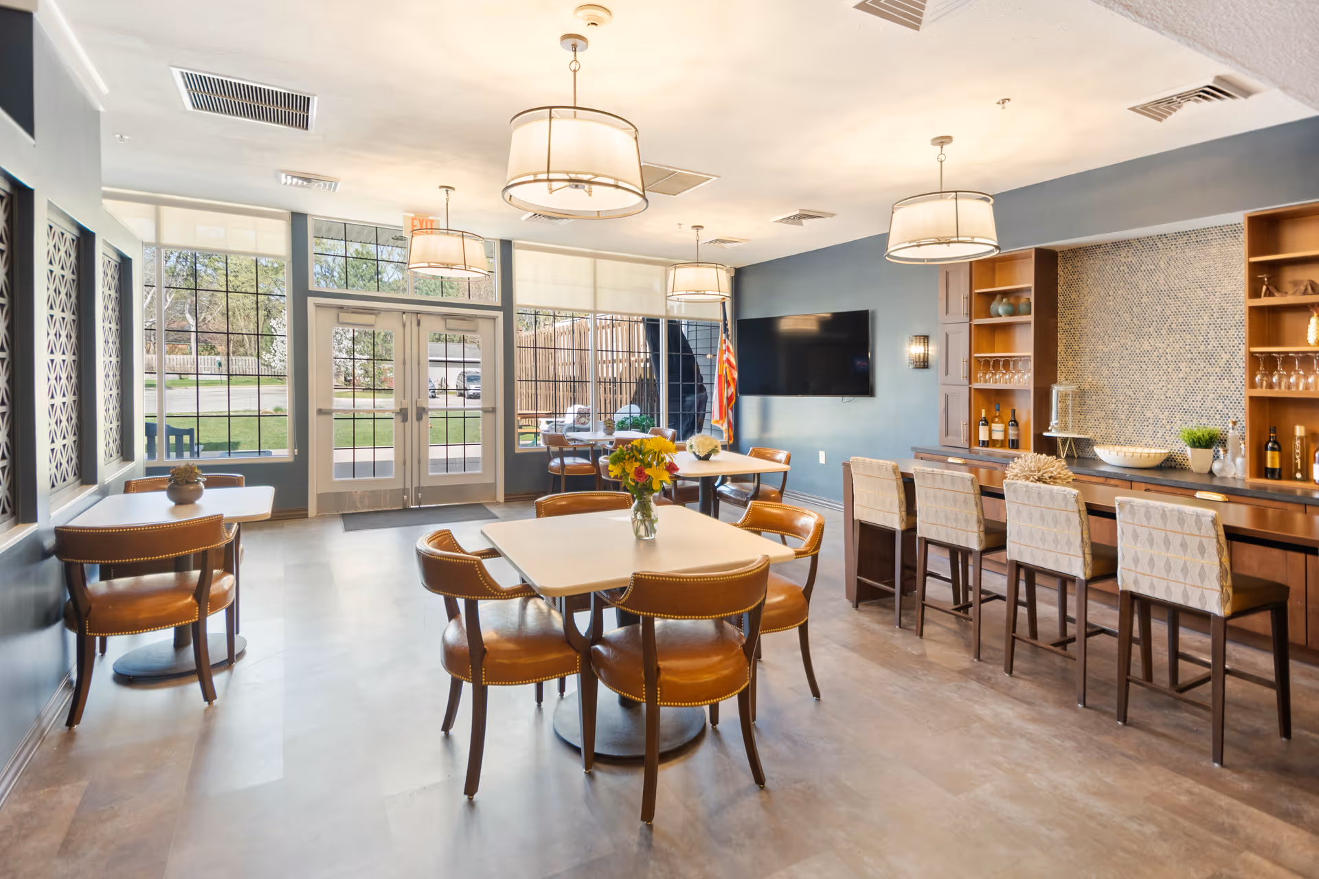 Bright communal dining and lounge area with tables and leather chairs, a bar counter with stools, wall-mounted TV, and large windows by the entrance.