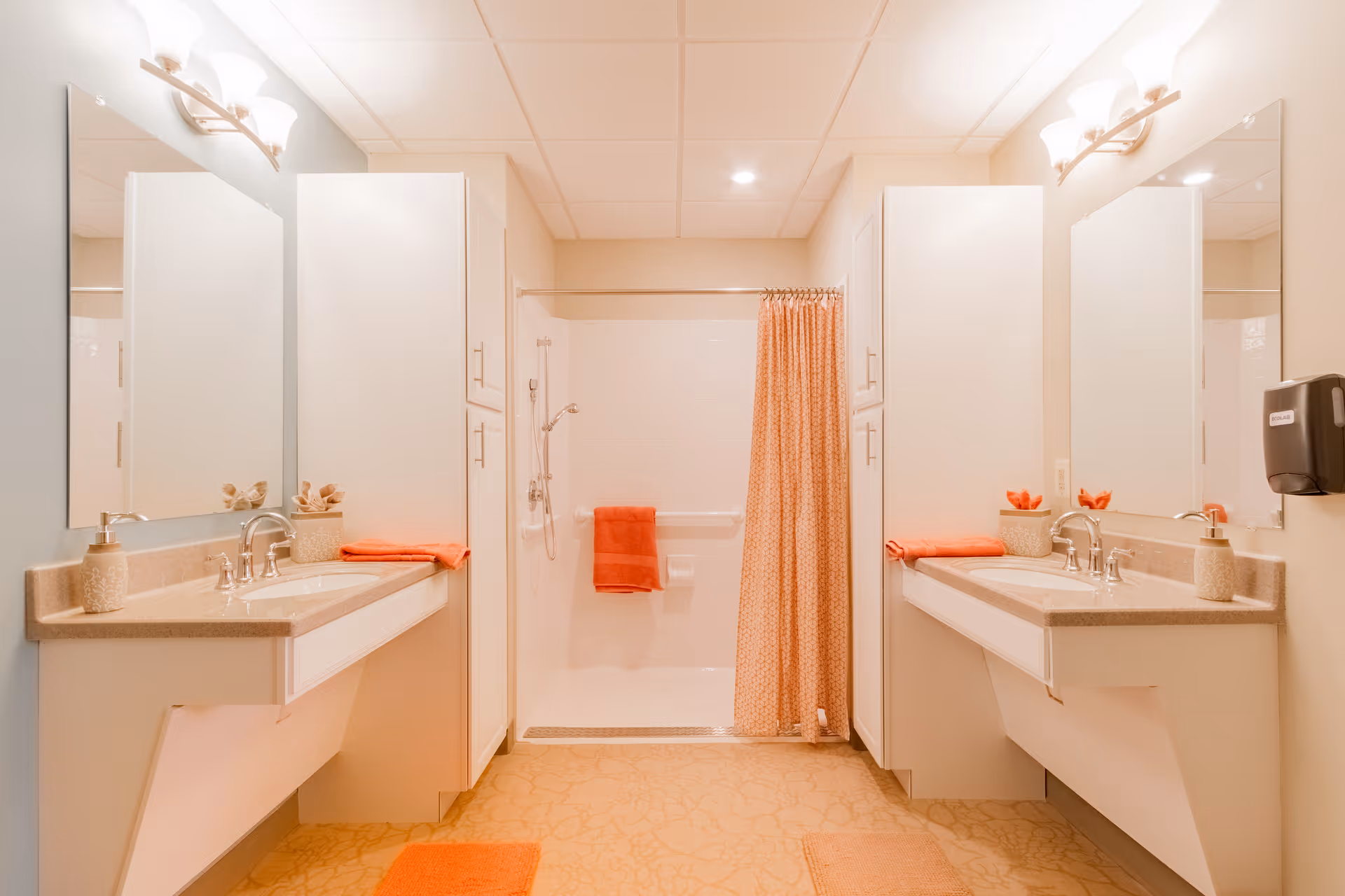 A bright, clean bathroom with two sinks and mirrors on either side, a walk-in shower with a patterned orange shower curtain, orange towels, and beige cabinets and countertops.