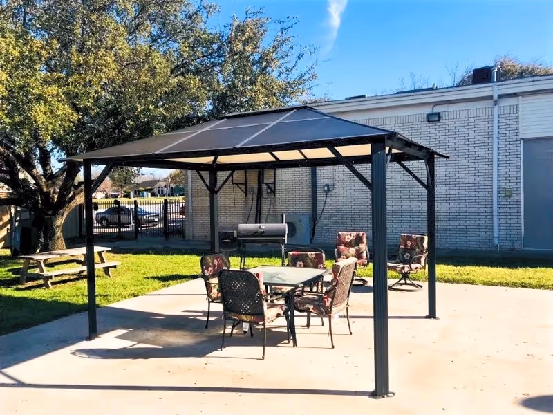 Outdoor patio area with a metal gazebo structure providing shade over a glass table surrounded by six cushioned chairs with floral patterns. In the background, there is a large tree, a picnic table, a barbecue grill, and a white brick building under a clear blue sky.