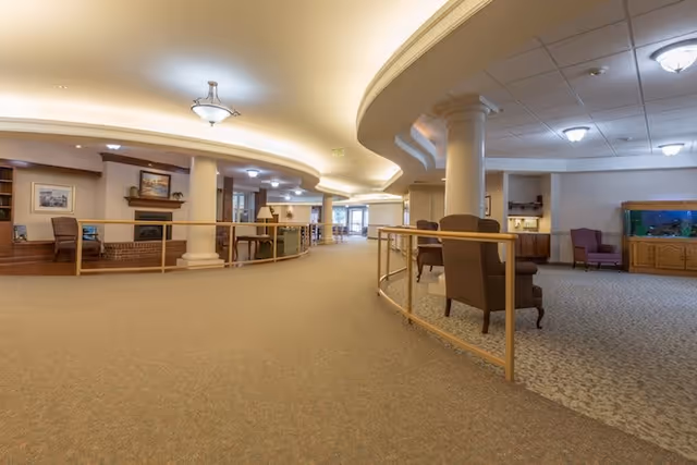 Spacious and well-lit interior common area of a senior living facility with beige carpeted floors, curved ceiling with recessed lighting, white columns, wooden railings, comfortable chairs, a fireplace, framed artwork, and a large fish tank in a wooden cabinet.