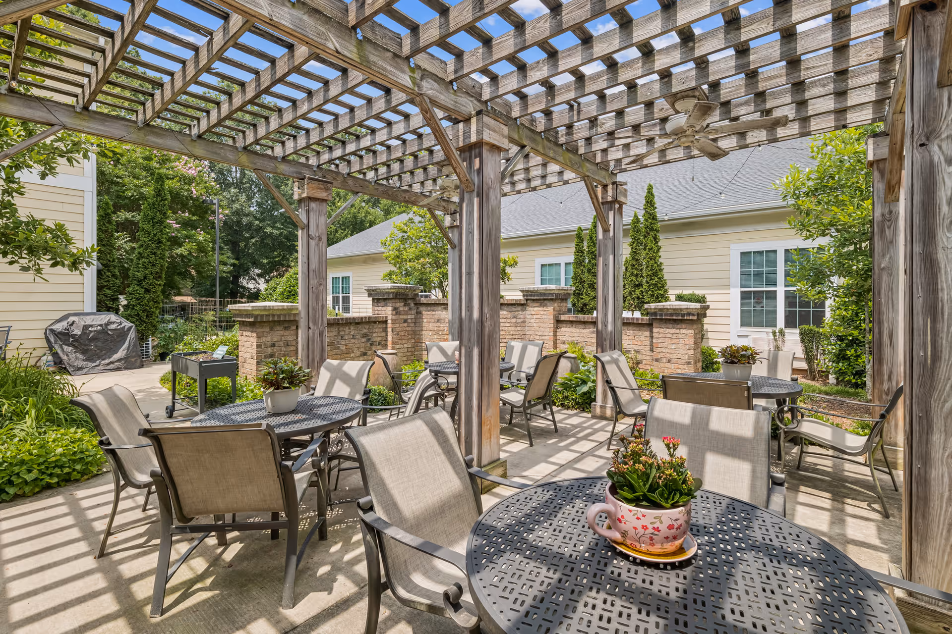Outdoor patio area at Cadence Huntersville featuring multiple metal tables with beige cushioned chairs under a wooden pergola. Potted plants decorate the tables, and there is greenery and a brick wall surrounding the space. A ceiling fan is attached to the pergola, and a covered grill is visible in the background.