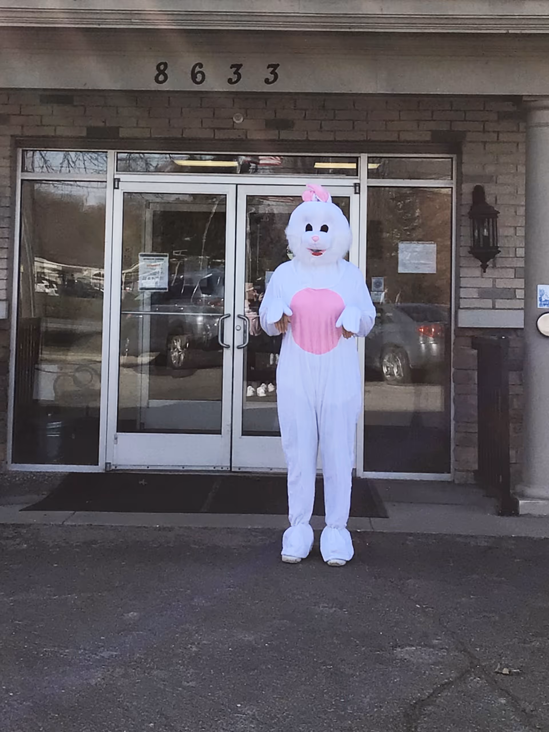 Person dressed in a white and pink bunny costume standing in front of the glass entrance doors of a building with the address number 8633 above the doors.