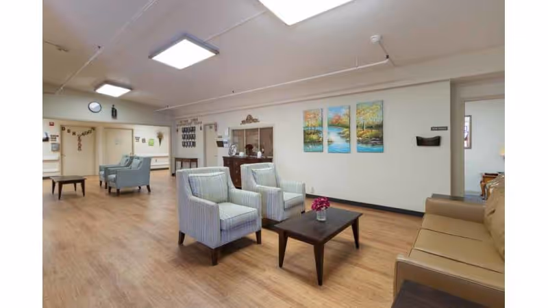 A spacious and well-lit common area in a senior living facility featuring two striped armchairs and a brown sofa arranged around a dark wooden coffee table with a small flower vase. The room has light wood flooring, white walls adorned with three colorful landscape paintings, and ceiling lights. Additional seating and a small table are visible in the background along with a hallway entrance.