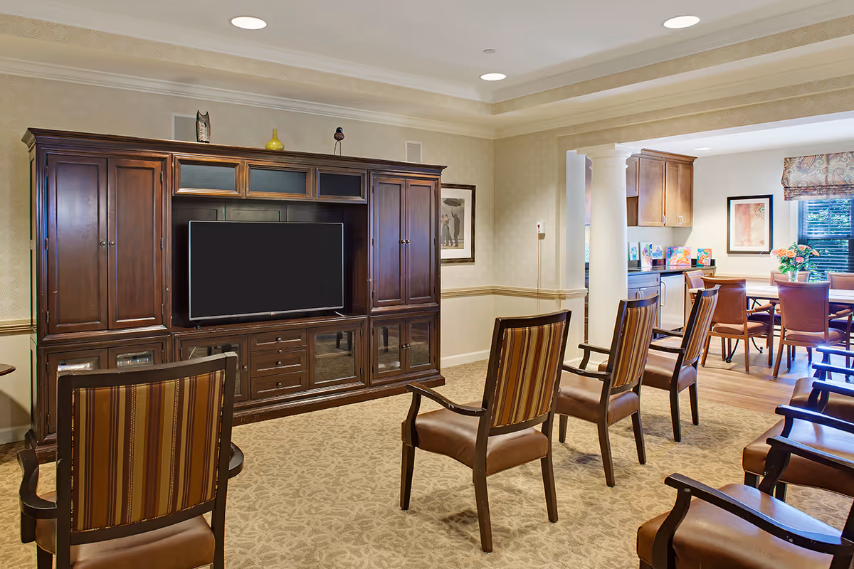 A cozy living room area with several wooden chairs featuring striped upholstery arranged facing a large wooden entertainment center with a flat-screen TV. The room has beige patterned carpet, wallpaper, and recessed lighting. In the background, there is a dining area with a wooden table, chairs, and a window with a patterned valance.
