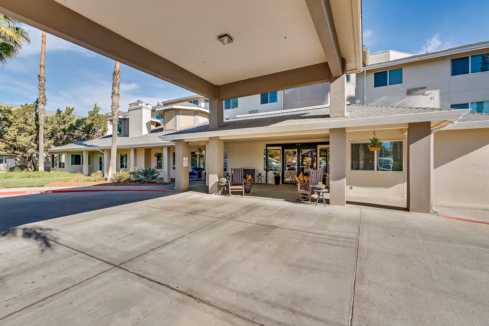 Entrance of The Remington Senior Living facility showing a covered driveway with two wooden rocking chairs and potted plants near the glass double doors. The building is light-colored with several windows and palm trees in the background under a clear blue sky.