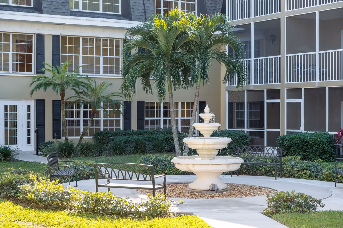 Courtyard featuring a three-tier fountain, benches, palm trees, and the surrounding senior living building with screened balconies.