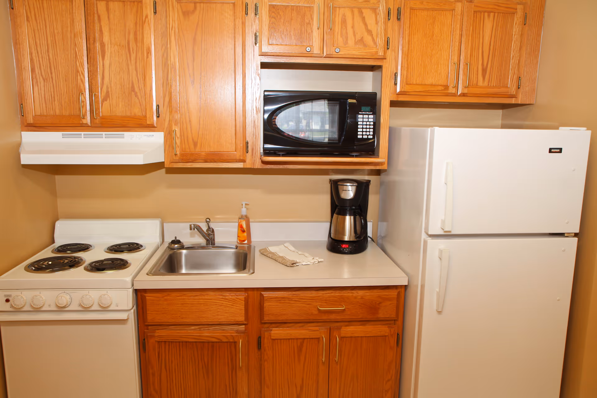 Compact kitchenette with oak cabinets, an electric stove, stainless sink, microwave, coffee maker, and a white refrigerator.