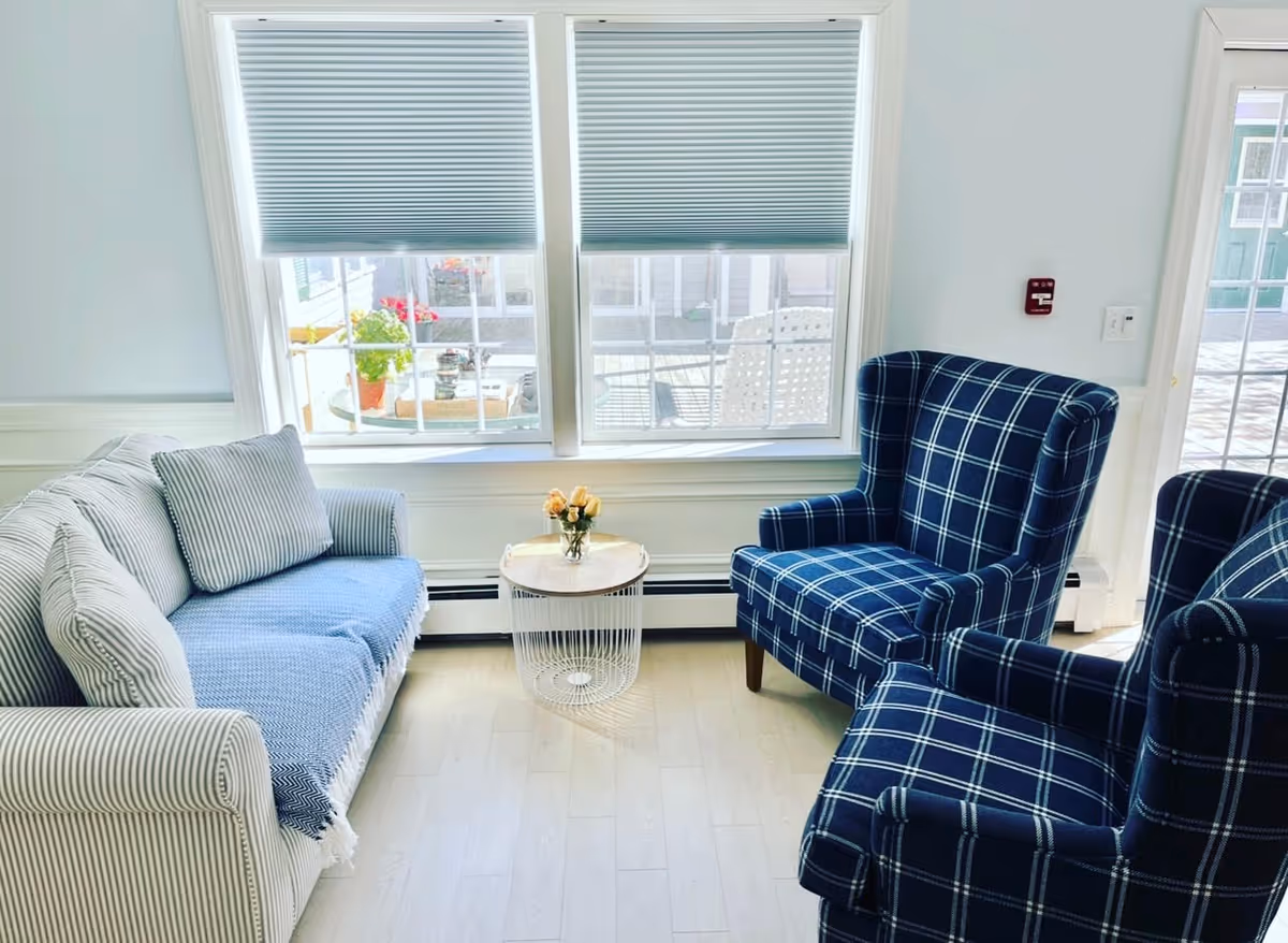 Sunlit sitting area with a striped sofa, two blue plaid armchairs, a small round table with flowers, and large windows.