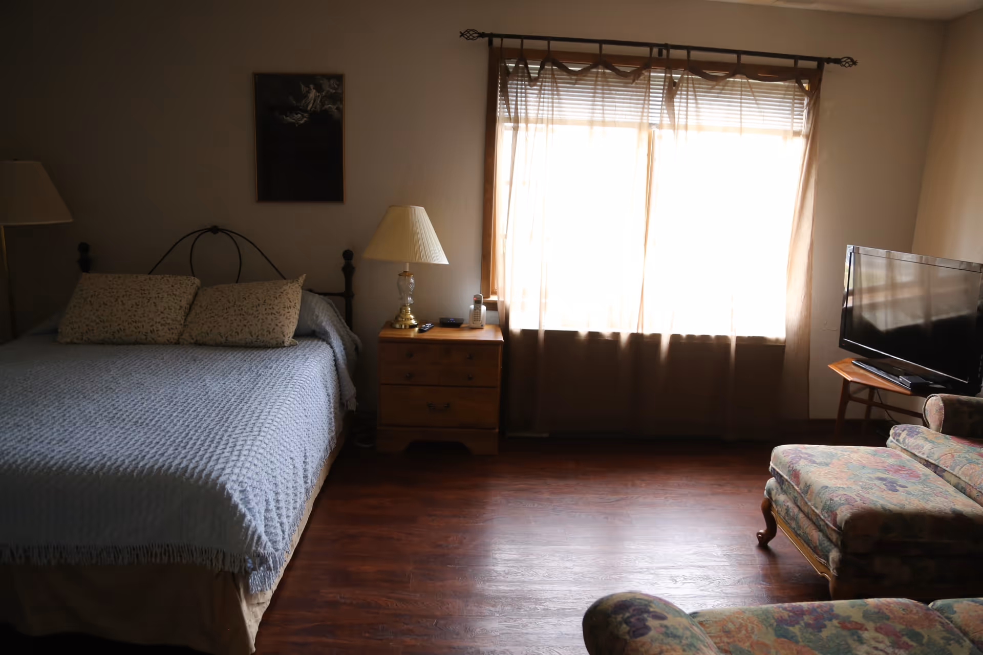 A cozy bedroom with a bed covered in a light blue textured blanket and two patterned pillows. Next to the bed is a wooden nightstand with a lamp, a cordless phone, and a remote control. A large window with sheer curtains lets in bright light. In the corner, there is a flat-screen TV on a small wooden table and a floral upholstered armchair with a matching ottoman. The floor is made of dark wood.
