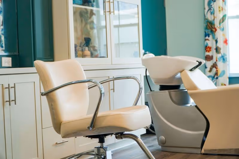 Interior view of a salon area with a beige salon chair and a white hair washing station. There are white cabinets with glass doors in the background and a window with colorful floral curtains.