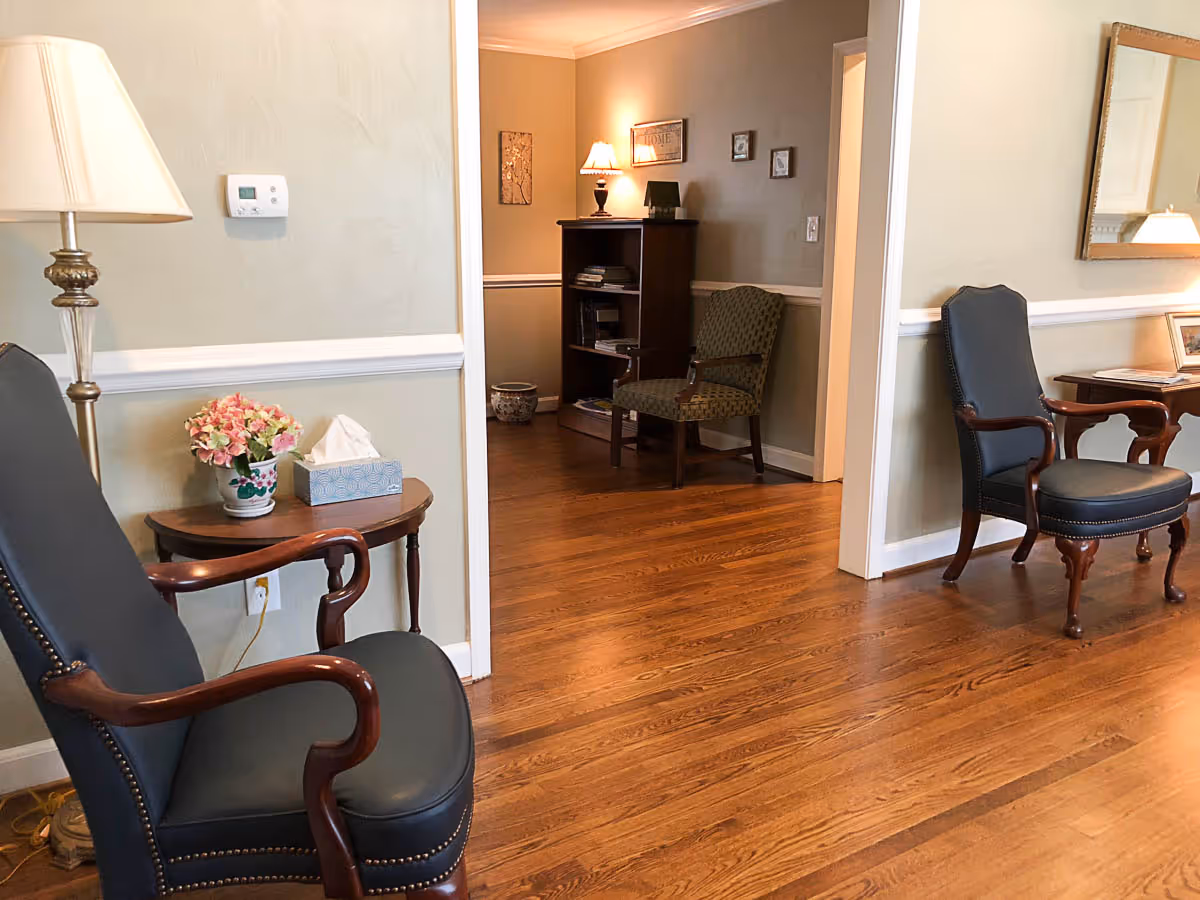 A cozy interior room with wooden flooring featuring two dark upholstered armchairs with wooden armrests, a small wooden side table with a flower pot and tissue box, a standing lamp, and a bookshelf with a lamp and framed pictures on the wall in the background.